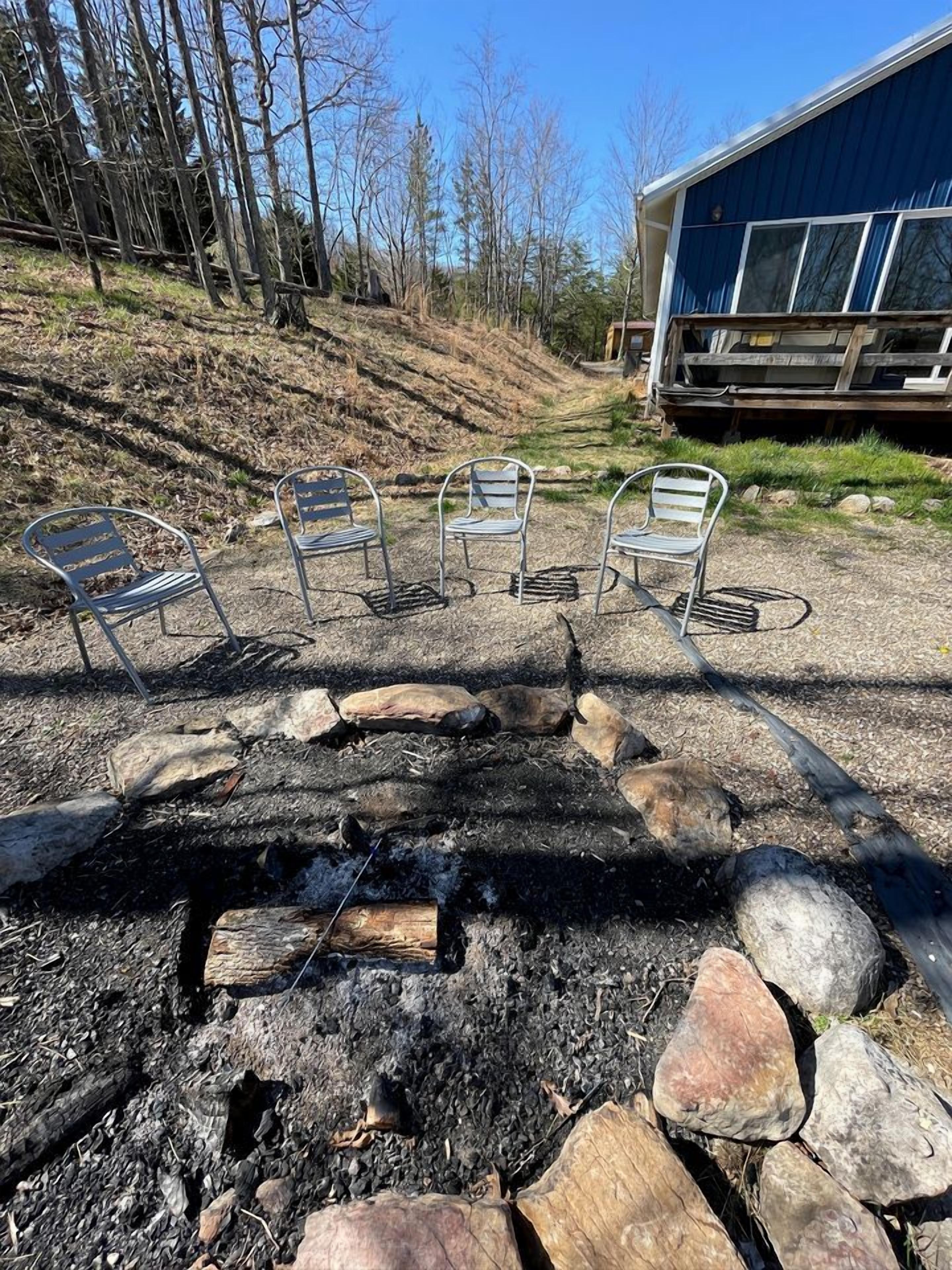 A fire pit with rocks and metal chairs in a backyard with a house and trees in the background.