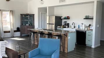 A spacious living area with a high ceiling, exposed beams, a central island with bar stools, a kitchen with modern appliances, and a cozy seating area with blue armchairs.
