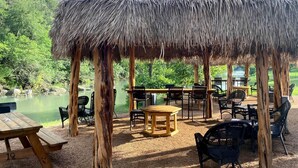 A thatched-roof pavilion with outdoor seating, a picnic table, and a view of a lake. - Glamping Adventure on the Guadalupe River: Cool Bell Tent near New Braunfels, Texas Perfect for All Ages (New Braunfels)