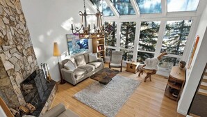 Een ruime woonkamer met een stenen openhaard, een groot raam met uitzicht op bomen, een kroonluchter en een gezellige zithoek.