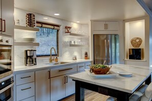 A modern kitchen with a large island, stainless steel appliances, and a view of a forest through the window.