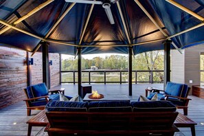 A covered outdoor seating area with a fire feature, wooden deck, and a view of a lake and trees.