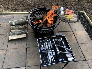 Un barbecue portable avec un foyer, une grille et un plateau contenant de la viande et des épis de maïs, le tout installé sur une terrasse.