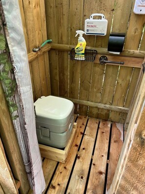 A wooden outdoor toilet with a grey container, a first aid kit, and a hand sanitizer dispenser.