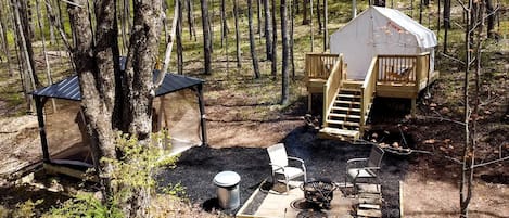 Un camping en el bosque con una pequeña construcción de madera, una mesa con sillas y un cubo de basura.