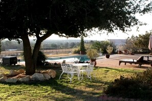 Um quintal com piscina, uma mesa e cadeiras brancas, uma espreguiçadeira e uma árvore grande.