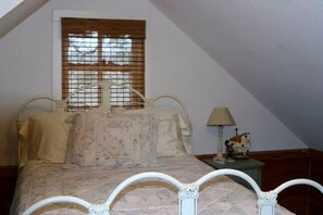 Ein Schlafzimmer mit Bett, Fenster mit Jalousien, Nachttisch mit Lampe und einem kleinen Dekorationsobjekt.