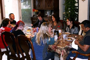 A group of people dining at a table with a floral tablecloth, wooden chairs, and a cabinet in the background.