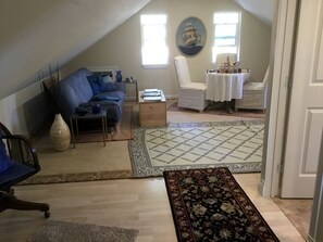 Ein Loft-Zimmer mit schrägem Dach, einem blauen Sofa, einem weißen Sessel, einem kleinen Tisch und einem gemusterten Teppich.
