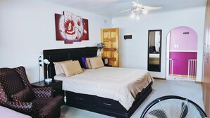 Un dormitorio con cama, mesita de noche, lámpara, silla, obra de arte, puerta, espejo y ventilador de techo.