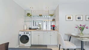 Une cuisine compacte équipée d’un lave-linge, un coin repas avec une table et des chaises, et un petit espace détente avec un banc.
