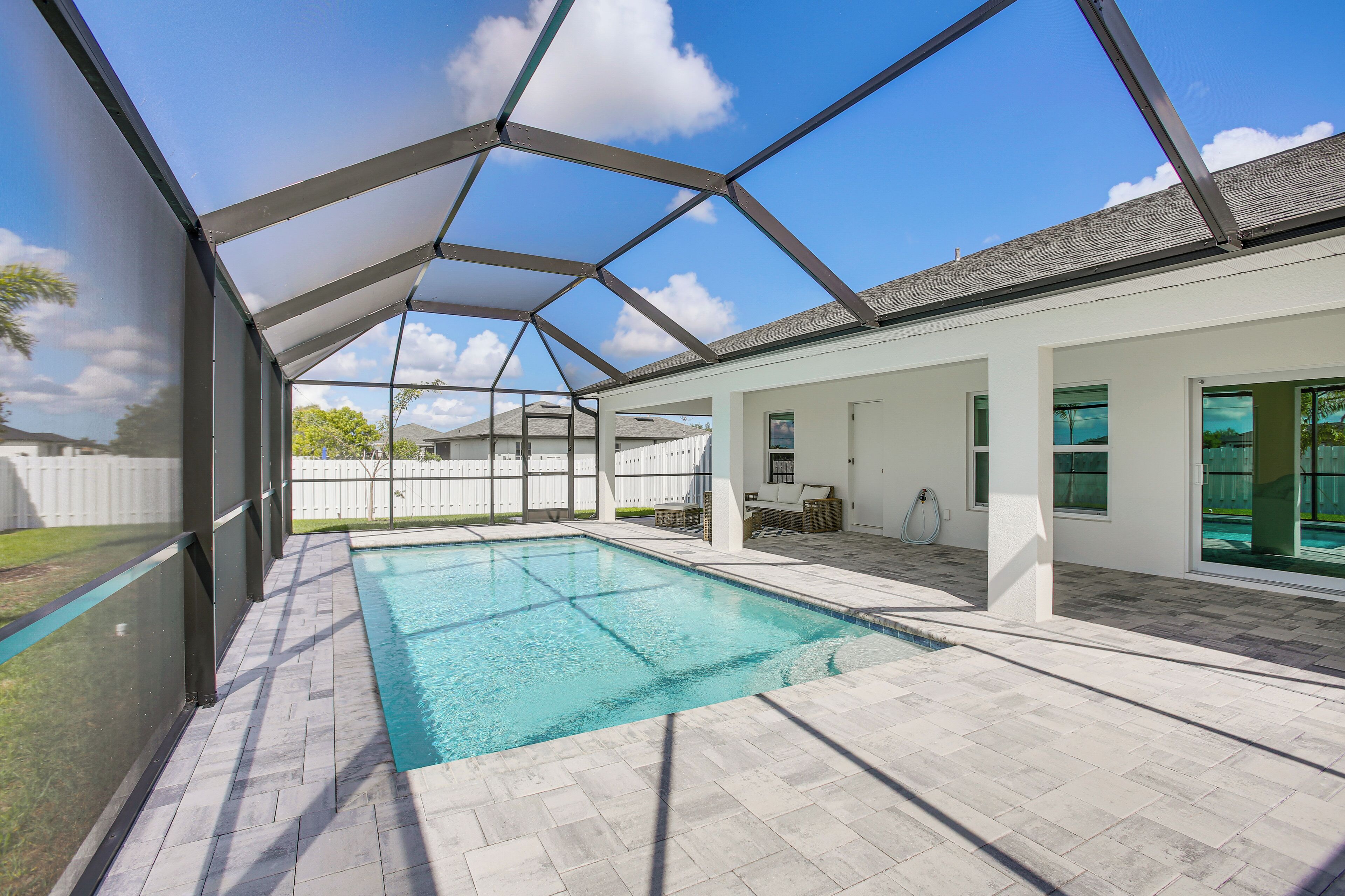 A swimming pool with a covered patio area, a white exterior wall, and a clear blue sky.