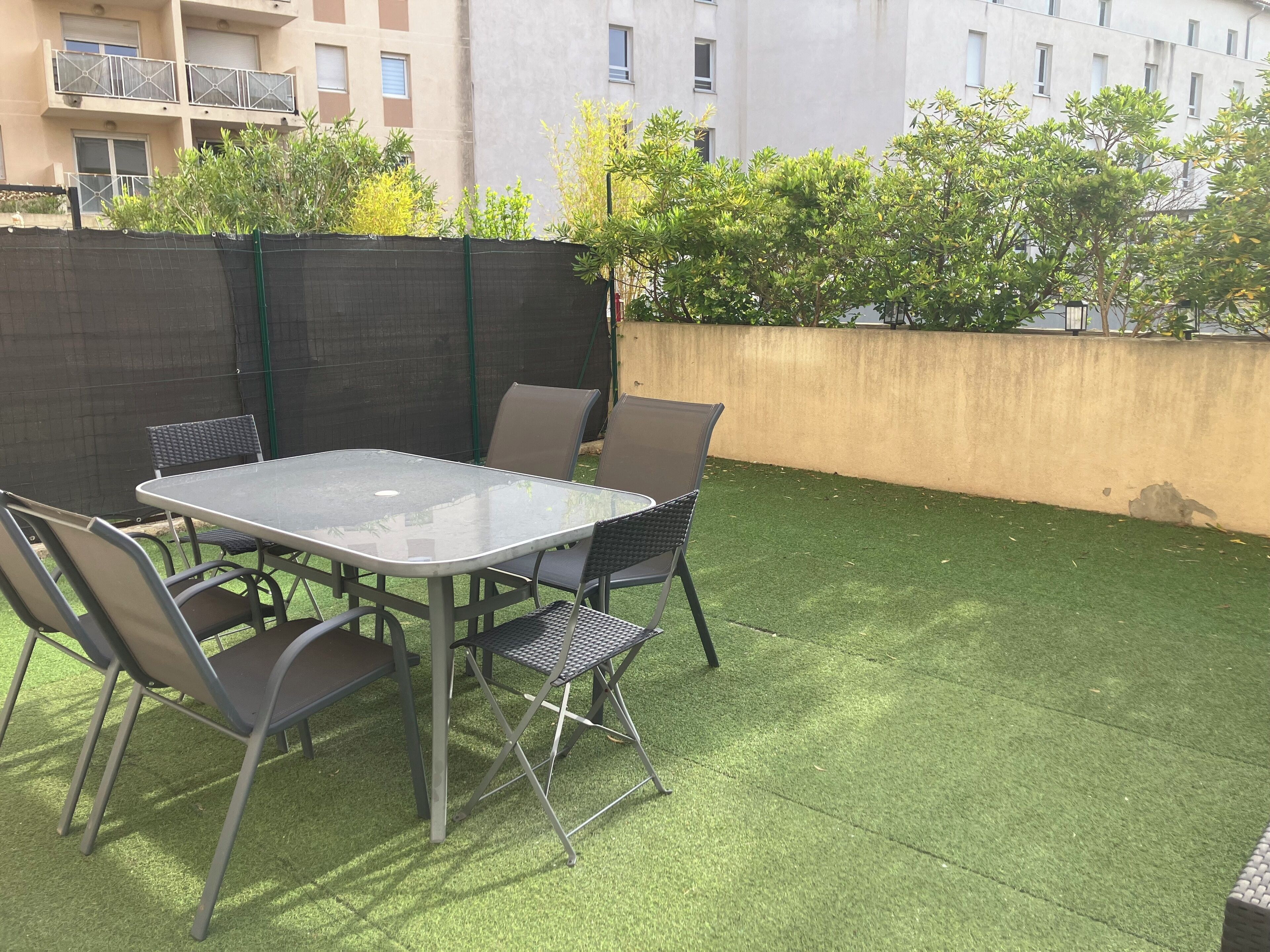 Une terrasse extérieure avec une table en verre et des chaises en métal, entourée de gazon synthétique et d’un petit muret.