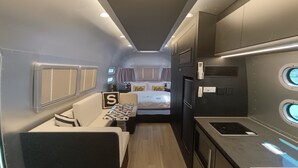 Interior de autocaravana compacto con kitchenette, cama con almohada que dice 'Hola' y pequeña zona de estar.