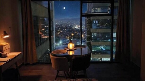 Una stanza con una scrivania, una sedia, un tavolo con una lampada, un'ampia finestra che si affaccia sulla città e un letto con copriletto decorato.