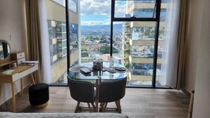 Una camera d'albergo con una grande finestra, una scrivania con una sedia, un letto e un poggiapiedi.