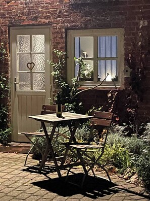 A brick building with a wooden door and window, a table with chairs, and greenery around.