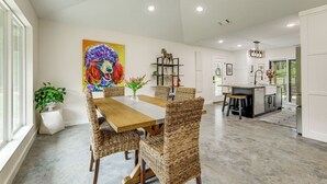 A spacious dining area with a wooden table and wicker chairs, a colorful painting on the wall, and a kitchen in the background. - Modern-Style Home w/ Patio, Near Greers Ferry Lake (Heber Springs)