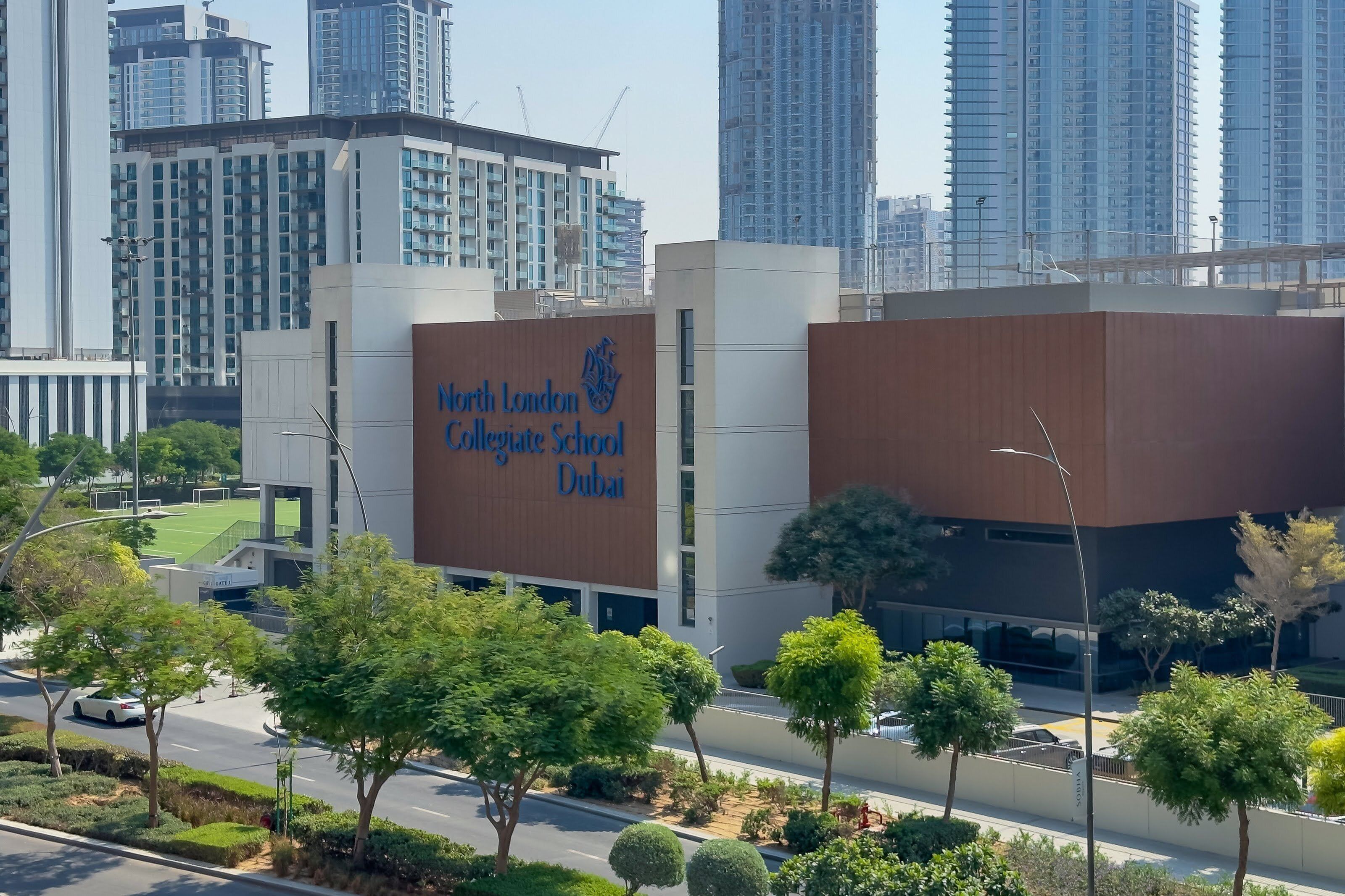 Um edifício moderno com a placa da North London Collegiate School Dubai, rodeado por vegetação e arranha-céus.
