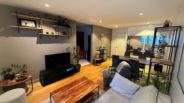 Ein modernes Wohnzimmer mit Flachbildfernseher, Couchtisch aus Holz, grauem Sofa und einem Essbereich mit dunklem Tisch und Stühlen.
