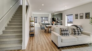 Een modern interieur met een trap, een zithoek met een bank en kussens, een eethoek met een tafel en stoelen, en een keuken met kasten en apparatuur.