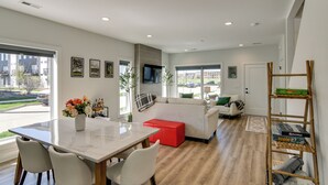 Een moderne woonkamer met een eethoek, een bank, een rode poef en een tv die aan de muur is bevestigd.