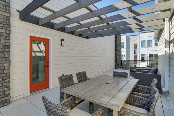 A covered patio with a wooden table and wicker chairs, a red door, and a view of neighboring buildings. - Rooftop Oasis: Downtown Des Moines Townhome (Des Moines)