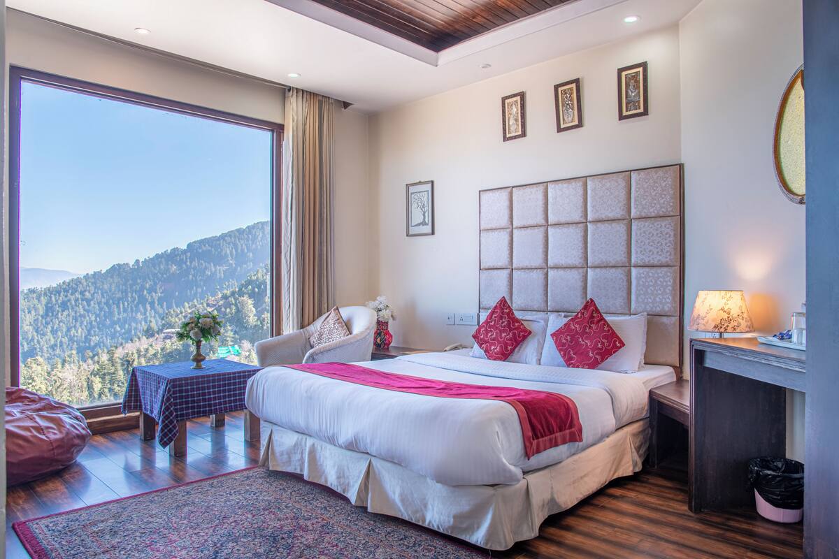 Un dormitorio con un ventanal grande, una cama con ropa de cama roja y blanca, una silla, una mesita con un florero y una mesita de noche con una lámpara.