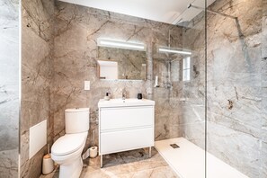 Un baño moderno con pared de mármol, cabina de ducha de vidrio, lavamanos blanco y un inodoro.