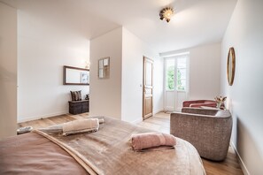A bedroom with a large bed, a small seating area, a dresser, and a door leading to another room.