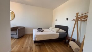 A bedroom with a bed, a chair, a wooden floor, and a wall-mounted light fixture.