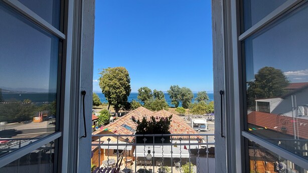 Vista pela janela: céu claro, um corpo d'água ao longe e uma rua arborizada.
