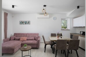 Ein modernes Wohnzimmer mit einem Ecksofa, einem Esstisch mit Stühlen, einem kleinen Beistelltisch mit einer Pflanze, Pendelleuchten, einer Klimaanlage, einer Uhr und einem Fenster mit Blick auf Bäume.