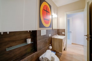 Un baño moderno con un mueble de tocador de madera, un lavamanos y un inodoro. Se ve un perchero con toallas y un rollo de papel higiénico.
