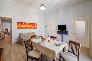 Un comedor con una mesa dispuesta para dos, sillas, un sof á, un televisor montado en la pared y una puerta.