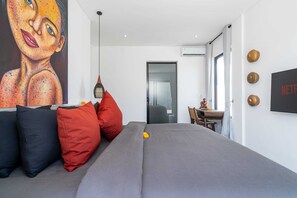 A modern bedroom with a grey sofa, red and black cushions, a hanging light fixture, a large painting, and a wall-mounted TV. - Villa Alexine (Seminyak)