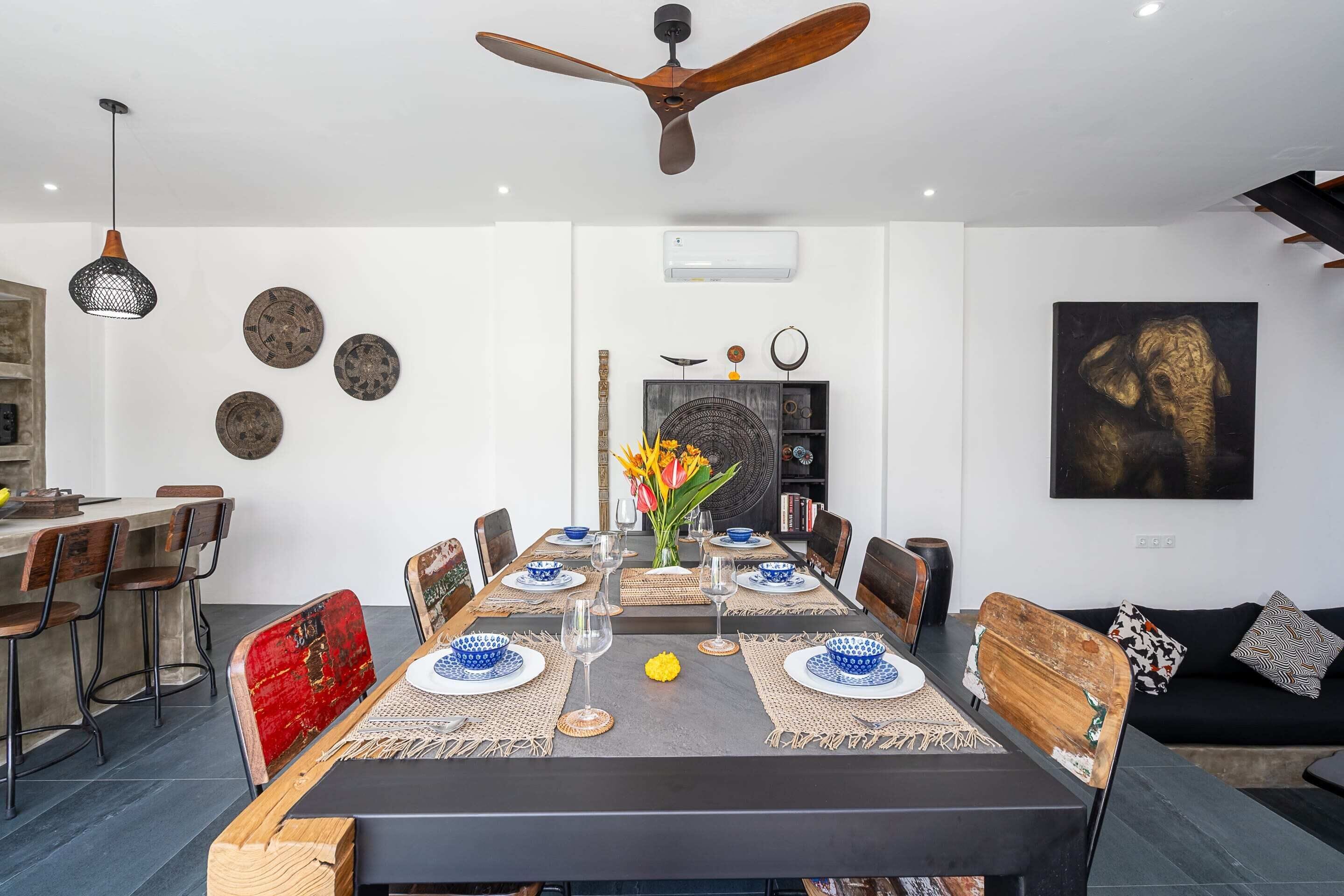 A dining area with a long table, chairs, a ceiling fan, wall decorations, and a sofa.