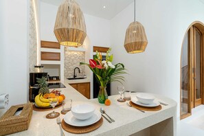 A modern kitchen with a granite countertop, hanging wicker pendant lights, a stone backsplash, and a dining area set with white plates and bowls. - Villa Atlas (Legian)