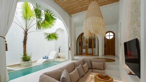 A modern living room with a large sectional sofa, a TV, and a pool area visible through an open archway. - Villa Atlas (Legian)