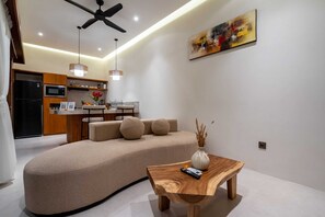 Ein modernes Wohnzimmer mit einem gebogenen Sofa, einem Holztisch, einem Deckenventilator und einer Küche im Hintergrund.
