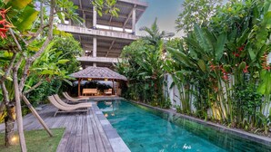 Ein Pool mit Liegestühlen, eine strohgedeckte Struktur und üppige tropische Vegetation.