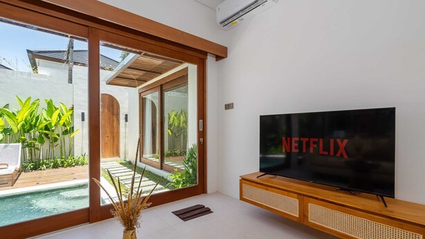 A modern living room with a large flat-screen TV, a wooden entertainment unit, and a sliding glass door leading to a pool area. - Villa Iris by Elysian (Kerobokan)