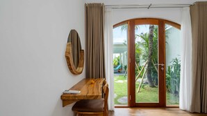 A room with a wooden desk, a chair, a round mirror, and a sliding glass door leading to a garden. - Villa Konstelasi (Tabanan)