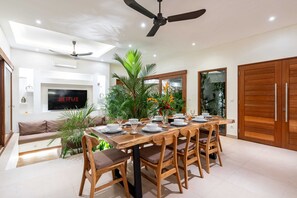 A dining area with a wooden table set for six, surrounded by chairs, a ceiling fan, and a TV displaying 'NETFLIX' on the wall. - Villa Naya Bali (Tabanan)