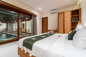 A hotel room with a bed, a desk, a chair, a large window, and a view of a pool. - Villa Eden by Elysian (Kerobokan)