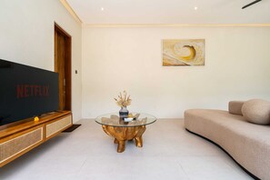 Een woonkamer met een flatscreen televisie, een glazen salontafel, een bank en een schilderij aan de muur.