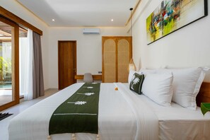 A hotel room with a bed, a desk, a chair, a window with curtains, and a painting on the wall. - Villa Luna by Elysian (Kerobokan)