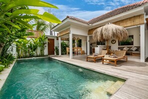 A poolside area with a wooden deck, lounge chairs, and a thatched umbrella. - Villa Sana (Tabanan)