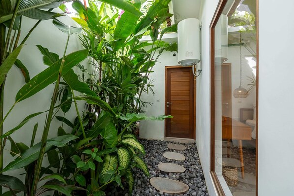 A narrow pathway with stepping stones leading to a wooden door, surrounded by lush green plants and a white wall. - Villa Sana (Tabanan)