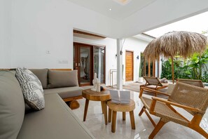A modern living room with a sofa, two wicker chairs, a round wooden table, and a thatched umbrella. - Villa Sana (Tabanan)